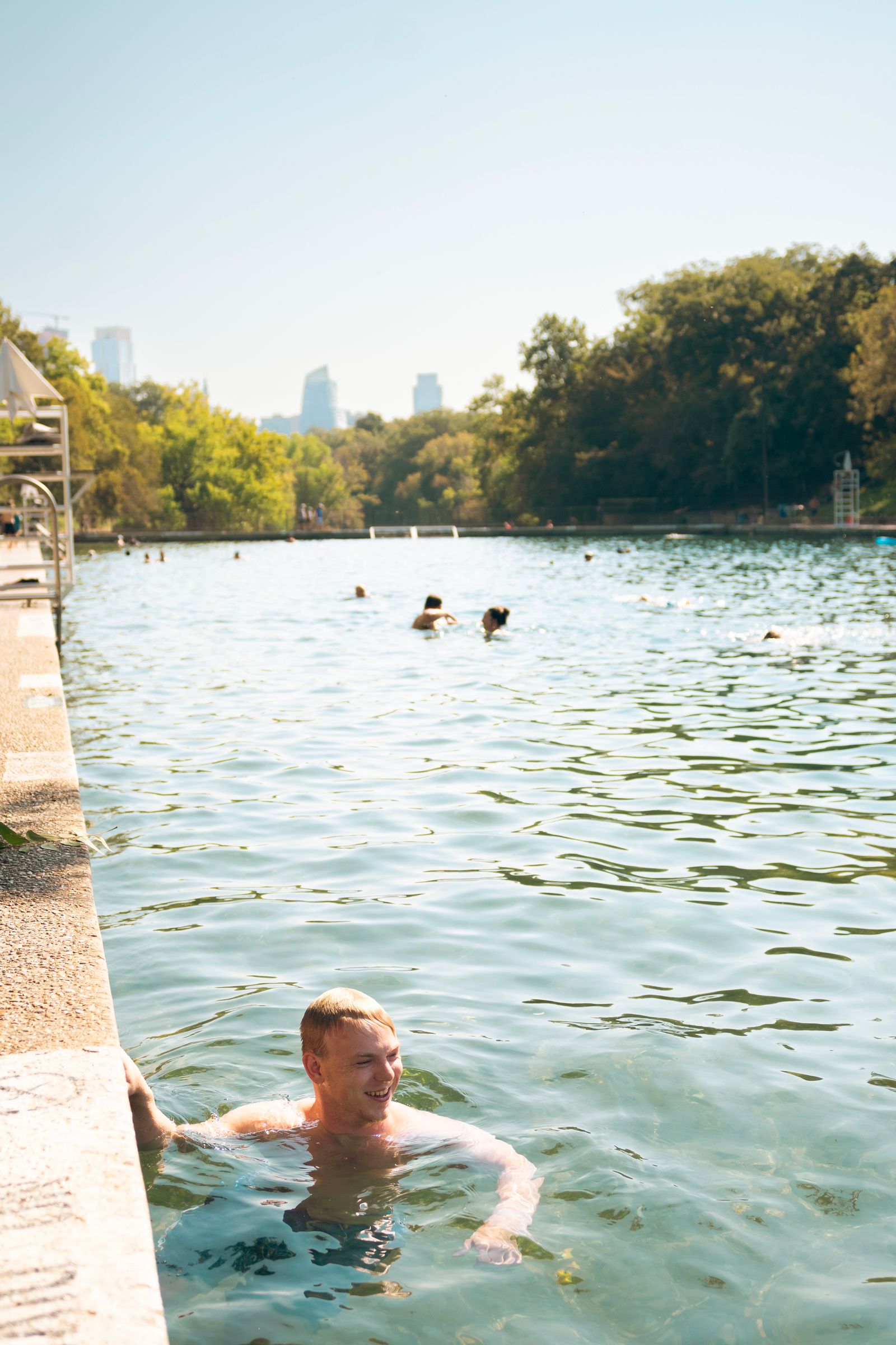 A man holding onto the ledge of the pool at Barton Springs, with the city in the background